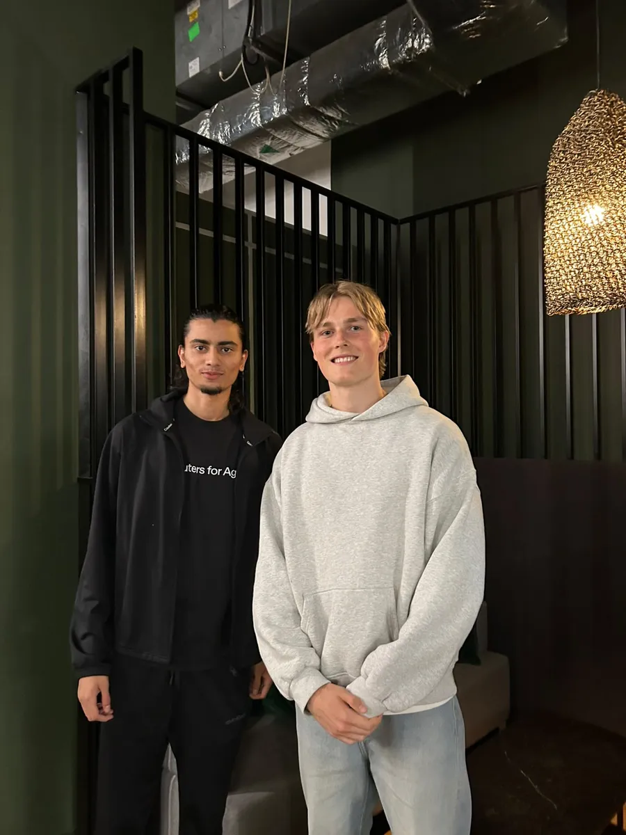 Rayhan and Samuel standing together in front of black slatted panels and a dark green wall