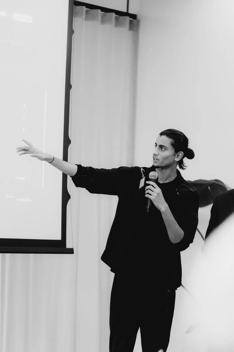 Black and white photo of Rayhan holding a microphone and pointing at a chart on the projected venue dashboard
