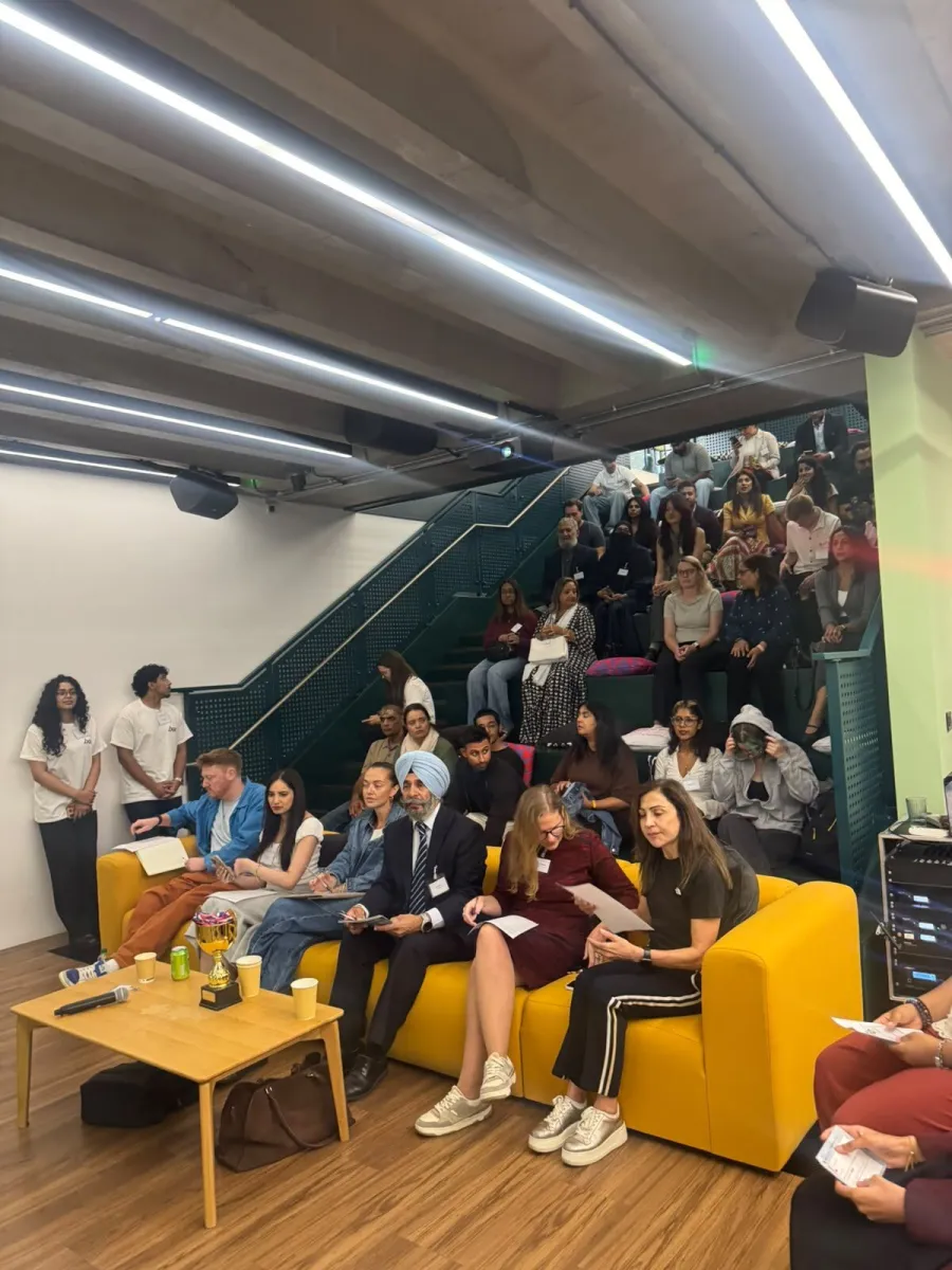 The judges on a yellow sofa with scoring sheets, a trophy on the table in front of them, and a full audience on tiered seating behind