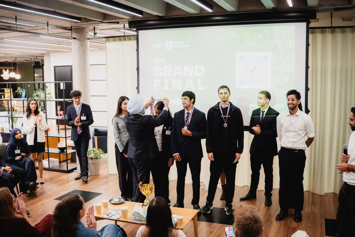 Jas Athwal putting a medal round a team member's neck in front of the Grand Final screen, with the rest of the Sidequest team already wearing theirs